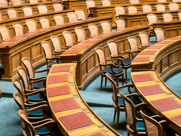 Vista interna de um plenário legislativo com bancadas de madeira e cadeiras clássicas organizadas em semicírculo, representando o ambiente de debates políticos e decisões regulatórias.